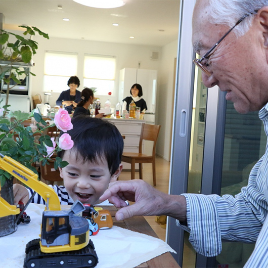 おじいちゃんと一緒に遊んでいる子どもと、キッチンで話をしている女性の写真