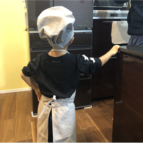 キッチンで帽子とエプロンを身に着けた子どもがオーブンを眺めている写真