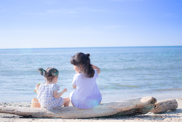 浜で流木に座っておやつを食べている姉妹の写真