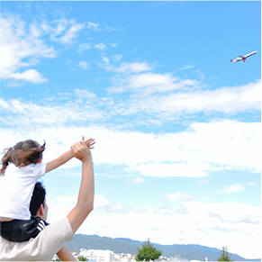 公園で空を飛んでいる飛行機に手を振っている大人と肩車している子どもの写真